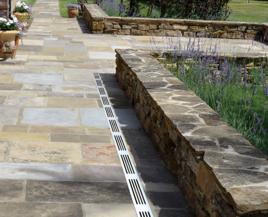 A long, low-profile metal linear drain installed along the edge of a stone patio next to a stone retaining wall and lavender plants.