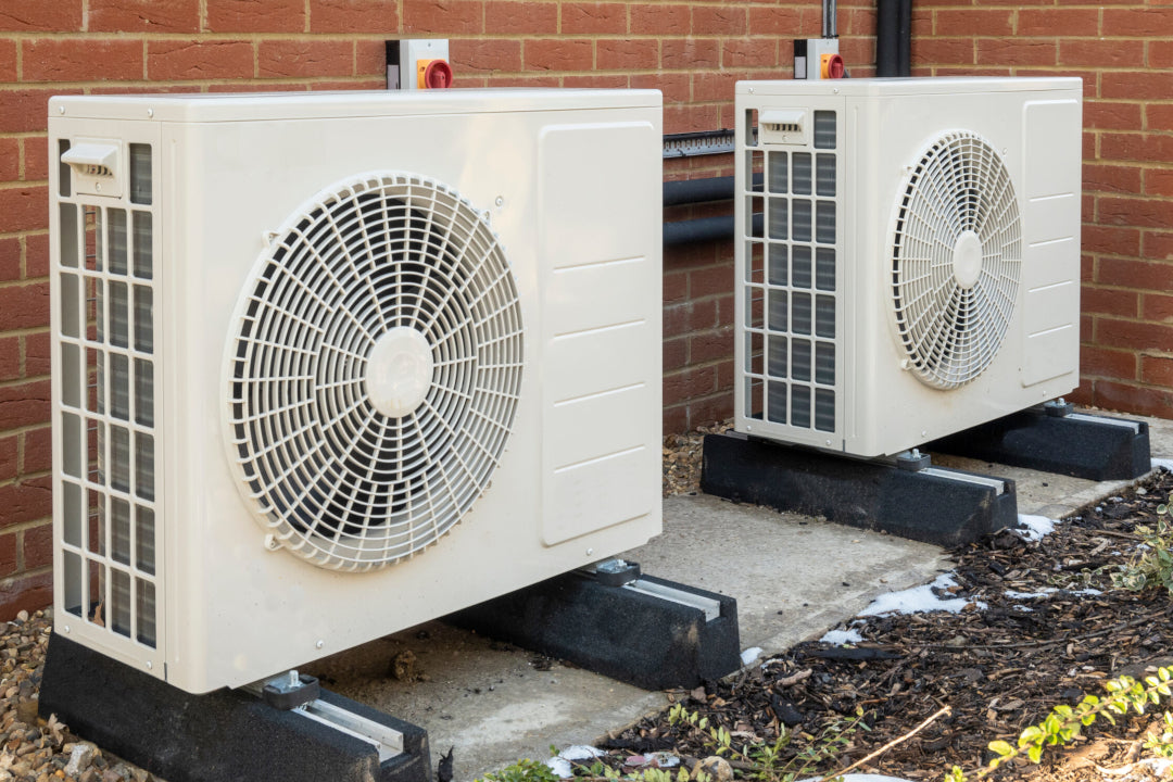 Two white outdoor heat pump or air conditioning units, positioned side-by-side against a brick wall, securely mounted on black rubber foundation blocks.