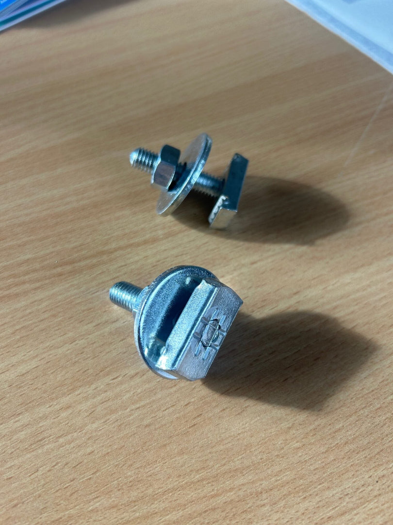 Two metal T-bolts with nuts and washers, designed for sliding into and securing equipment to the aluminum track of the mounting bases, resting on a wooden surface.